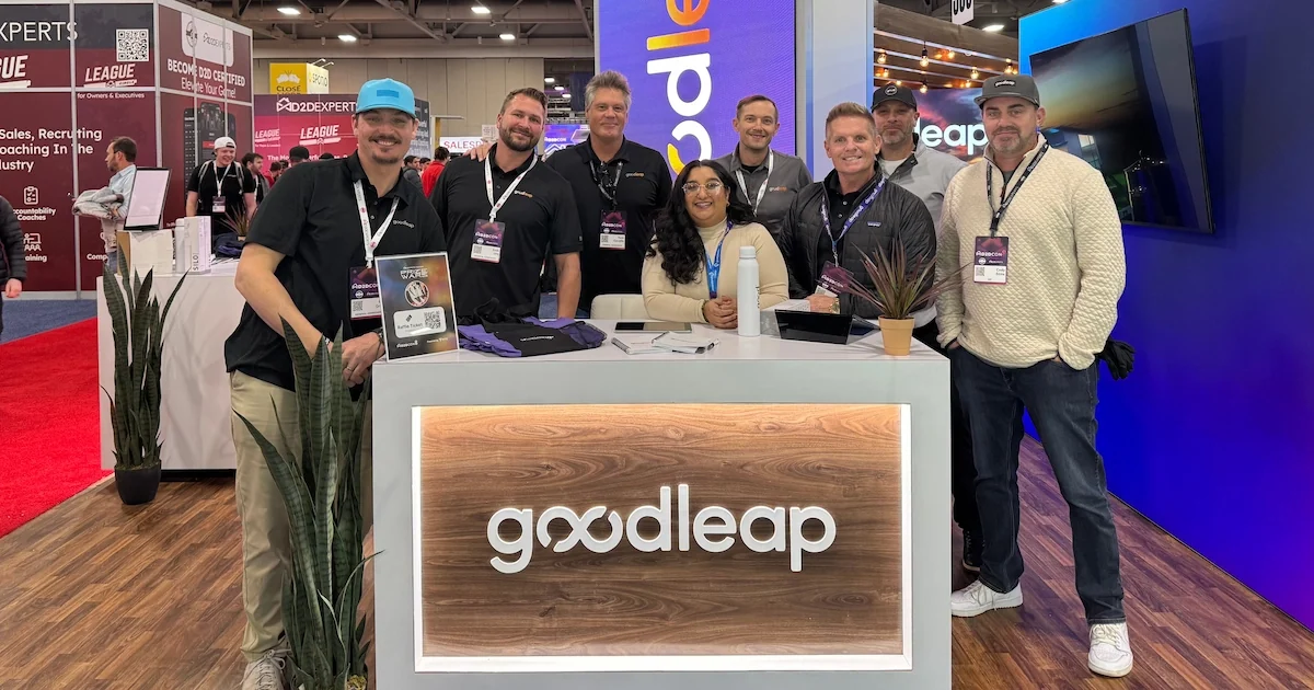 A group of GoodLeap team members poses at their trade show booth, featuring a sleek white counter with the illuminated GoodLeap logo. The backdrop includes a digital display, branding elements, and a busy expo floor with industry booths and attendees.