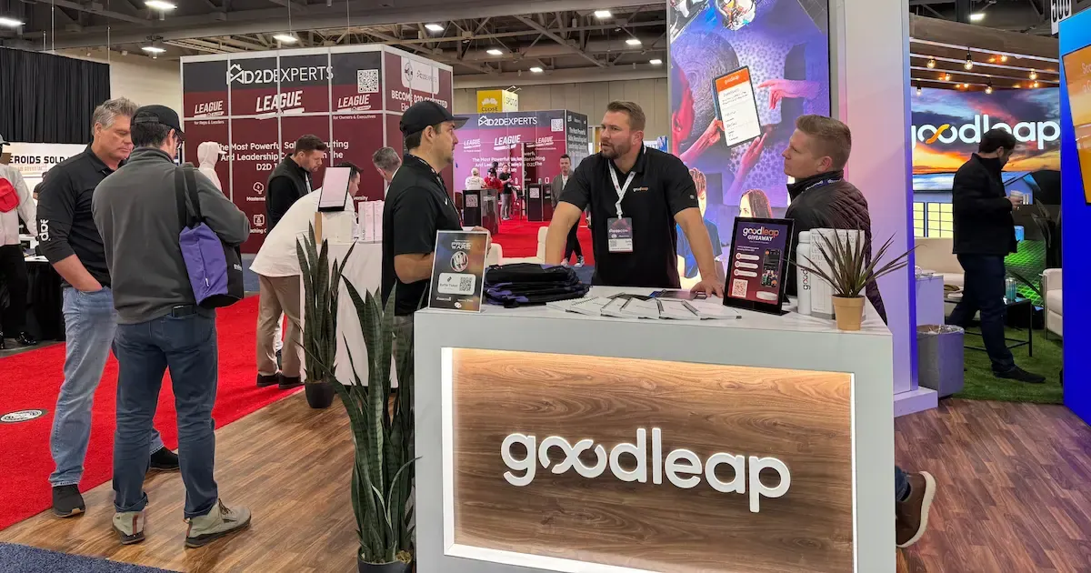 GoodLeap team members engage with attendees at their trade show booth, featuring a sleek white counter with an illuminated logo. The booth includes promotional materials, digital displays, and branding, with a busy expo floor in the background.