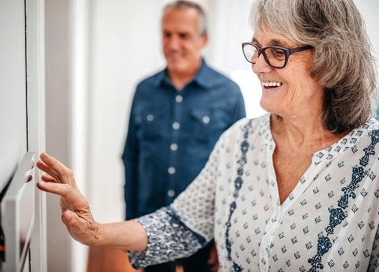 Older woman adjusting a smart thermostat with a man standing behind her.