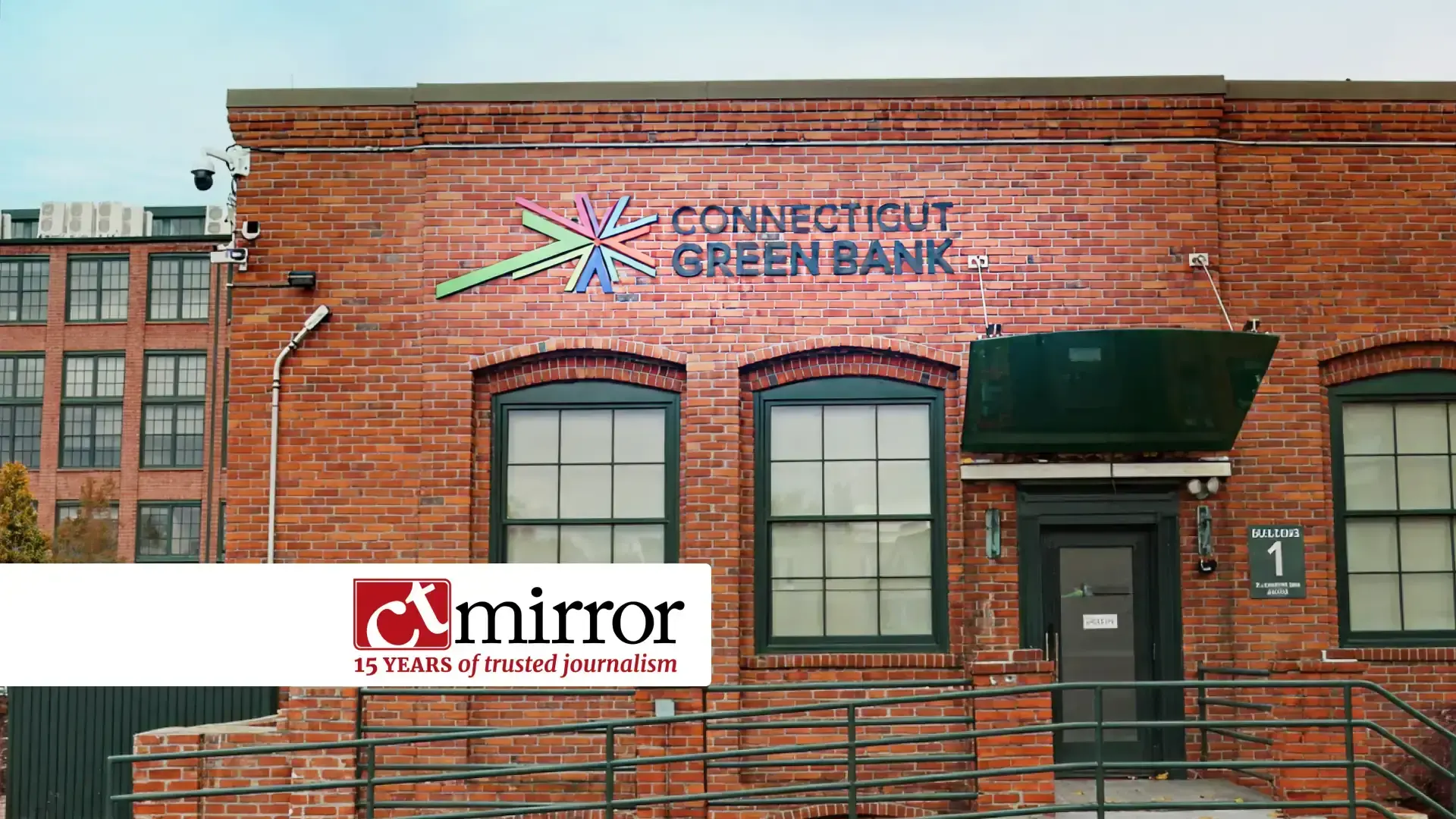 Exterior of the Connecticut Green Bank building with a red brick facade, two large windows, and the organization’s colorful logo above. A green awning covers the entrance door, and a CT Mirror banner in the foreground notes ‘15 years of trusted journalism.’