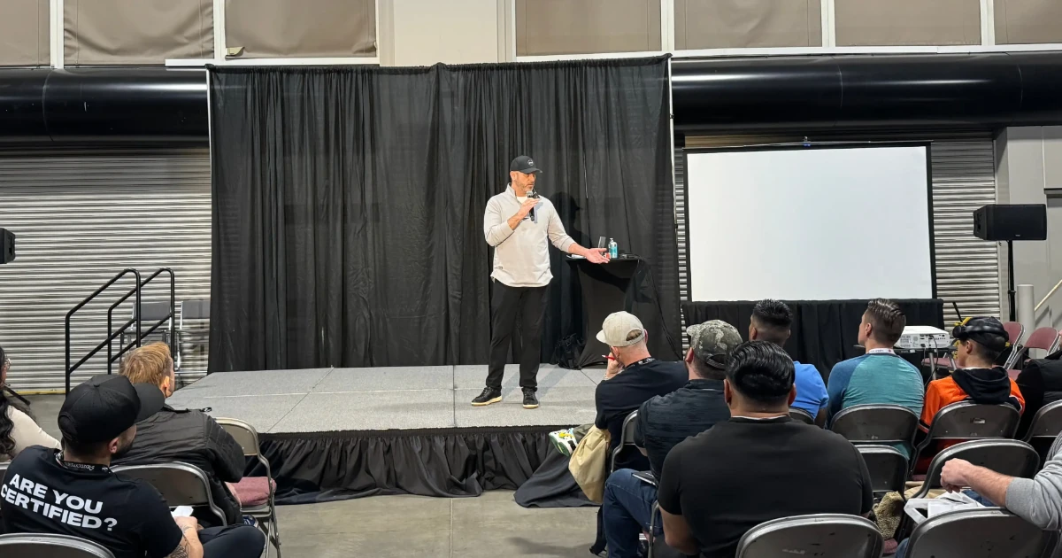 A speaker presents on stage at a trade show, engaging an audience seated in rows. The setup includes a black curtain backdrop, a projector screen, and a podium with water and notes. Attendees listen attentively in a professional expo setting.
