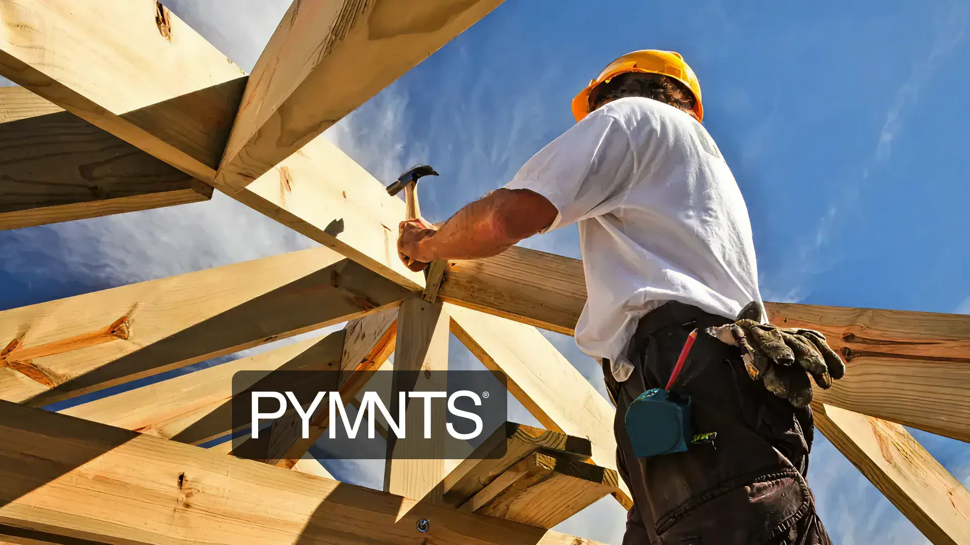A construction worker wearing a yellow hard hat and tool belt is hammering a nail into a wooden roof frame under a clear blue sky. The worker is viewed from a low angle, emphasizing the structure of the wooden beams. A “PYMNTS” logo is overlaid on the bottom left of the image.