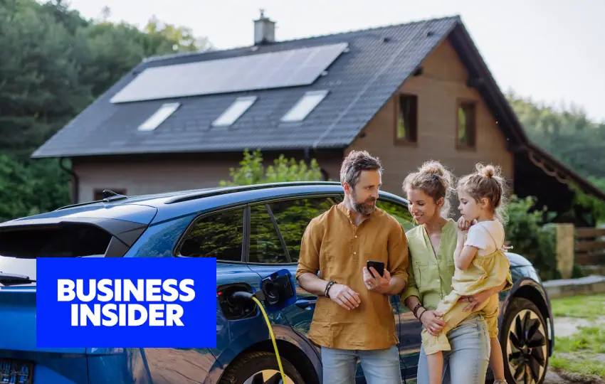 Family standing beside an electric vehicle charging at home with rooftop solar panels visible, alongside a “Business Insider” label.