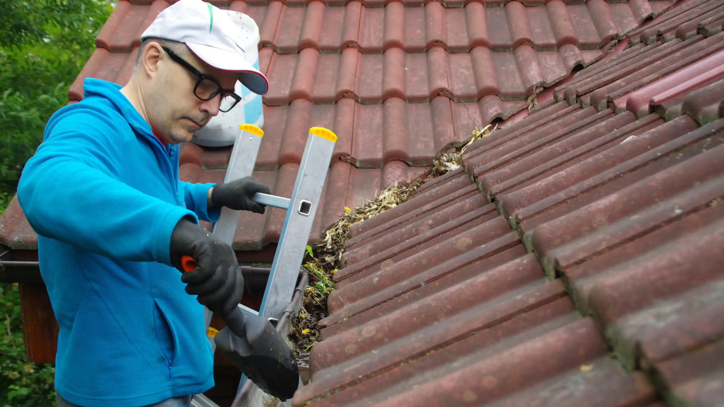 A homeowner on a ladder cleaning leaves from their gutter