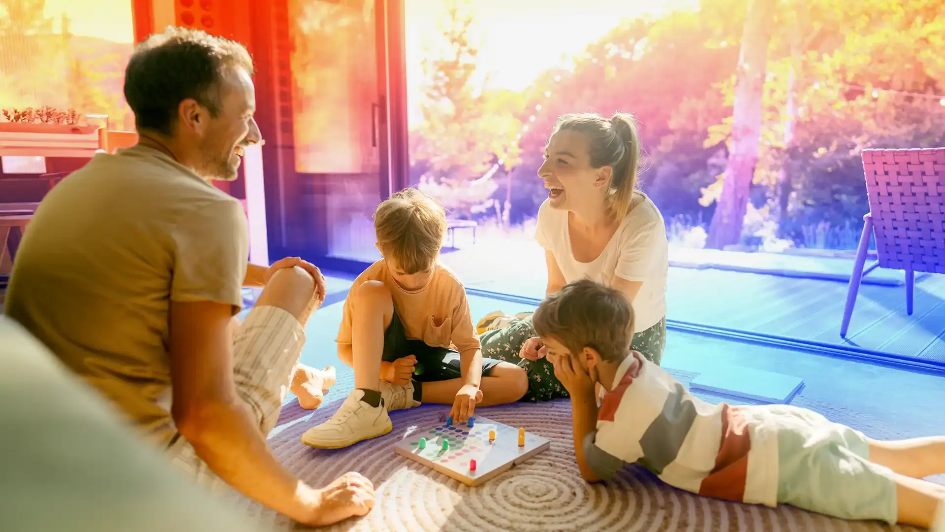 Family of four sitting on a rug in a sunlit room, happily playing a board game together.